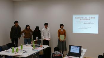 壁に映し出された資料の左側に立っている5名の参加者が発表を行っている様子の写真