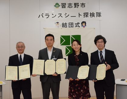 後ろに壁に「バランスシート探検隊結団式」と書かれた紙の前で、4名の関係者がそれぞれ協定書を持って一列に並んでいる集合写真