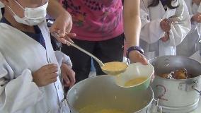 ニンジンを使って作られたスープを給食当番の児童が先生と一緒に給食の配膳で準備している写真