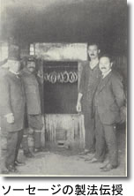 奥にソーセージが吊るされている手前左右にソーセージの製法伝授された男性が2名ずつ並んでいる白黒写真