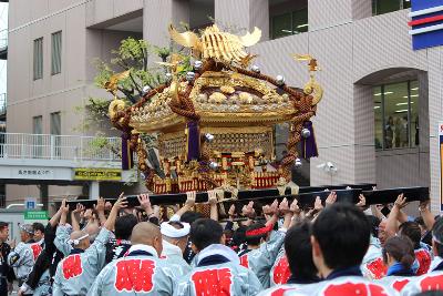 背中に藤と書かれた法被を着た方々が神輿を担いで市役所仮庁舎前を通っている様子の写真