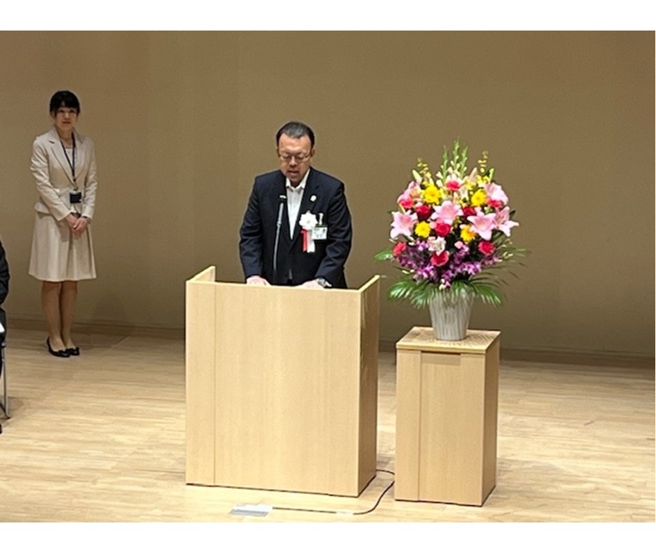 日の丸や習志野市の紋章がステージ正面に掲げてあり、ステージの演台前に立ち開式の言葉を述べている小熊副学長の写真