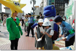 黄色の被り物に緑のスーツの上着を着た人の横で子供達が婦警さんにキャンペーンの案内をもらっている写真