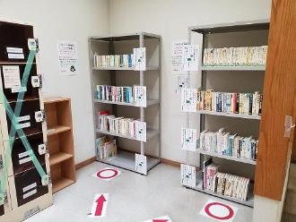 谷津図書館内の部屋の壁側に2つの本棚が設置され、床に矢印と大きな〇(丸)印が貼られている写真
