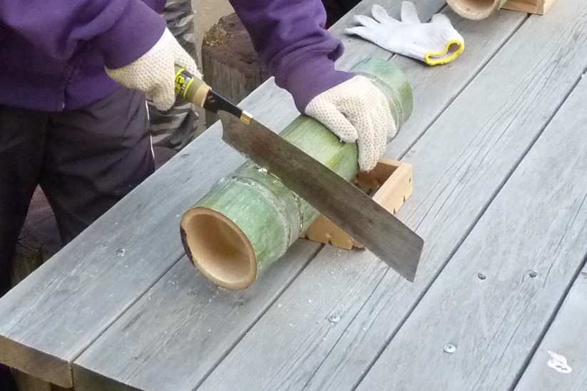 屋外テーブルの上で30cm程度に切られた竹にのこぎりで切れ目を入れているところ