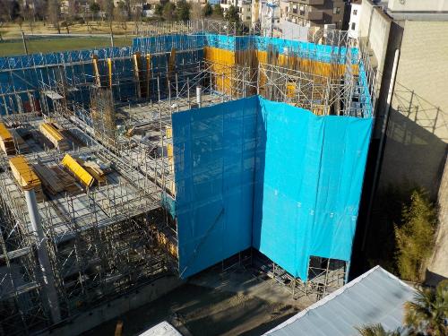 建物の周囲を足場が組まれて水色のシートで囲まれている建設途中の中層階の部分の写真