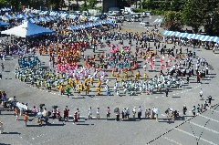 祭りの大きな会場で、カラフルな色の衣装を着た参加者が輪になって踊っている様子を上空から撮影した写真