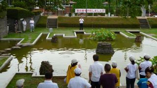 人工池の奥に宮本市長が立って話しをしているのを、池を挟んた手前に立っている参加者が聞いている写真