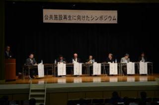 ステージの上に「公共施設再生に向けたシンポジウム」と書かれた幕が吊られ、ステージ上に7名のパネラーが横一列に並んで座っている写真
