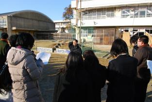 小学校の校舎と体育館の手前に立ち男性が話しをしているのを参加者が聞いている写真