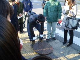 男性が下水道のマンホールの蓋を上げているのを参加者が見ている写真