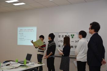 壁に映し出された資料の右側に立っている5名の参加者が発表を行っている様子の写真