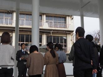 複合施設の建物の軒下で、スーツ姿の男性が話ているのを参加者が聞いている後ろ姿の写真