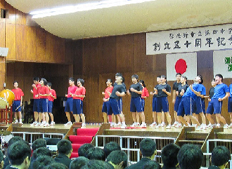舞台の上で左から赤い体操服を着た生徒達、真ん中に黒い体操服を着た生徒達、右に青い体操服を着た生徒達が手を腰に当てエール交換をしている写真