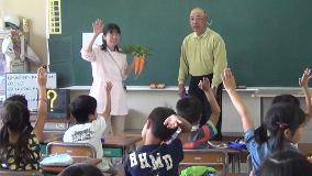 黒板の前に1人の男性と左手に2本のニンジンを持って右手を挙げている1人の女性が立ち、席に座っている児童たちも手を挙げている授業中の写真