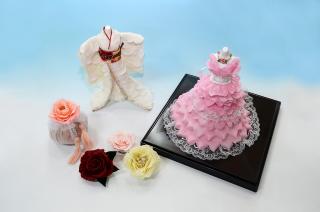 ピンク色のドレスや白い着物の工芸品と、赤、黄色、ピンクのバラが置かれてある写真