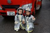 消防車の前で子供用の防火服のグッズを着てピースしている二人の子供の写真