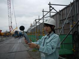 足場を組んだ工事現場で騒音計を持ち仕事をする海和 春香さんの写真