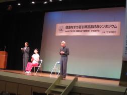 舞台上で椅子に腰かけている天井澤愛里沙氏と立ち上がっている青山敏彦氏、左端で手話通訳をしている女性の写真
