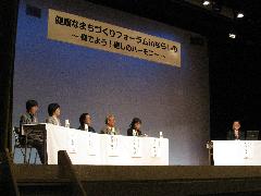 舞台上に設置されたテーブルの席について討論しているシンポジウム参加者の写真
