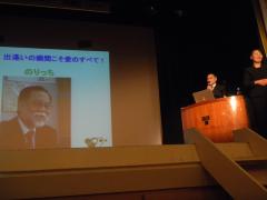 資料が映し出された舞台上のスクリーンと演壇で講演している島内憲夫氏の写真