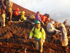 富士山8合目でたくさんの登山者の方が座っている近くで松岡秀善さんがご来光に照らされ笑顔でカメラを見ている写真
