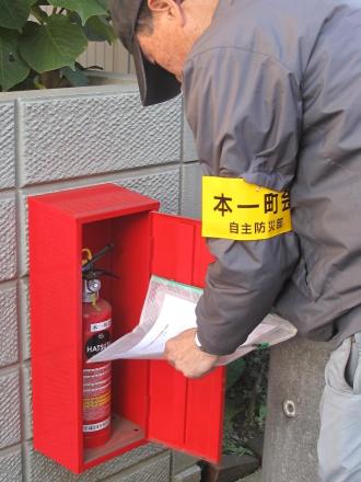 作業着を着た男性が赤いボックスに入った消火器を塀に設置している写真