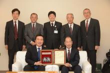 椅子に座った宮本市長と長谷川さんが賞状と盾を持ち、その後方に関係者の方が立ち記念撮影している写真