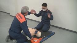 両手を上げて離れている救急隊員の間に横たわっているパッドを貼った傷病者のマネキンに電気ショックを行っている様子の写真