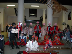 着物を着た数名の女性が琴の前に座り、後方に法被を着て頭にピンク色の被り物をした人々が立ち、演奏をしている様子の写真