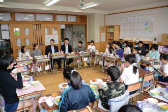 教室の机を円の字に並べ、給食を食べている津田沼小学校の子ども達や大人の写真