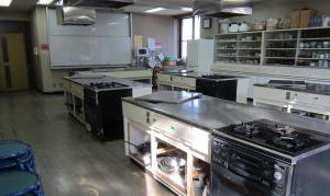 前方に白板があり、右側の壁には鍋や調理器具が収納されている棚、2口のガスコンロが設置されている調理台が4台並んでいる調理室の写真