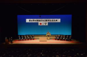 ステージ上の中心に演台があり、演題の両側に6名ずつ関係者が座っている記念式典の様子を写した写真