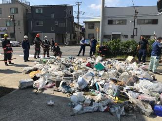 沢山のゴミが一角に広げて置かれて、職員の方々や消防服を着た方々がゴミの周りに集まっている様子の写真