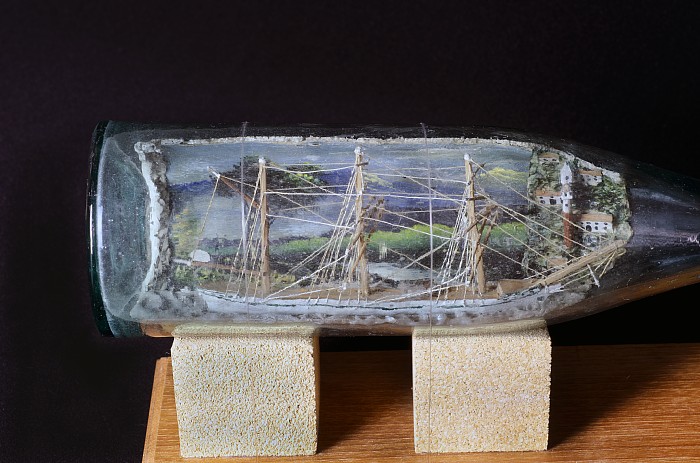 透明のボトルの中に船が作られ、背景には家々や木々が描かれているボトルシップ1例目の近接写真
