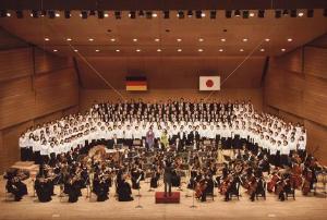 舞台の前方で指揮者に合わせて楽器を演奏している人達、後方で黒と白の衣装を着た人たちが合唱をしている習志野第九演奏会の様子の写真