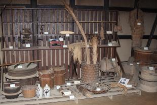 木製の蓋が付いた窯のような物、竹ざる、桶、お神酒入れなどの民具が展示されている写真