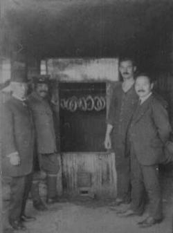 ソーセージが吊るされた釜、その前に立っている飯田技師とカール・ヤーン、ソーセージの製法を伝授された男性2名の白黒写真