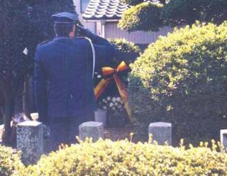 墓地の前で制服を着た男性が頭の横に右手をかざす敬礼をしている写真