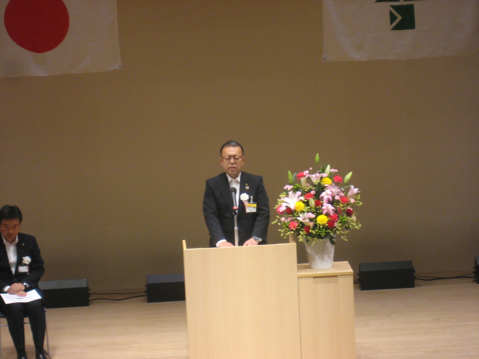 日の丸や習志野市の紋章がステージ正面に掲げてあり、ステージの演台前に立ち開式の言葉を述べている小熊副学長の写真