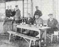 飲み物が置かれた長机の席に3名の男性が座り、その後方に数名の男性が立っている白黒写真