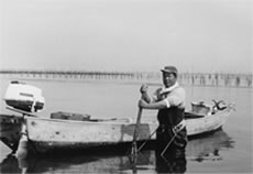 帽子を被った男性が船を背に写っている白黒写真