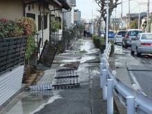住宅のフェンスが歩道に倒れ、歩道には液状化した水が溜まっている液状化被害の写真