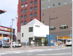 レンガ色の建物の前に配置された、四角の建物に三角屋根をした白い外壁の津田沼駅前交番の建物全体を写した写真