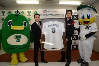 額に入った福浦選手のユニフォームを一緒に持っている宮本市長と山室球団社長、その横に立っているナラシドとマー君の写真