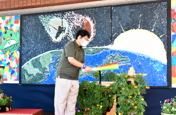 壁に飾られた大きなちぎり絵の前に立ち点火棒でランタンへ火をつけている久松さんの写真