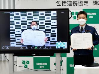 協定書を開いて見せているモニター画面の田中理事とモニター画面の横に立って協定書を見せている宮本市長の写真