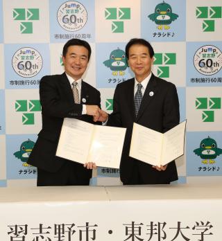左手に協定書を持ち、右手で握手をしている山崎学長と宮本市長の記念写真