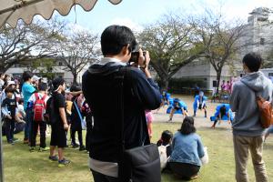 園庭で踊りを踊っている子供達を男性がカメラで写しているところを後方から写した写真