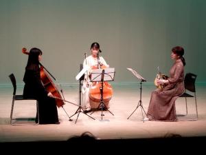 舞台の上で3名の女性が2台のチェロとホルンで演奏をしている写真